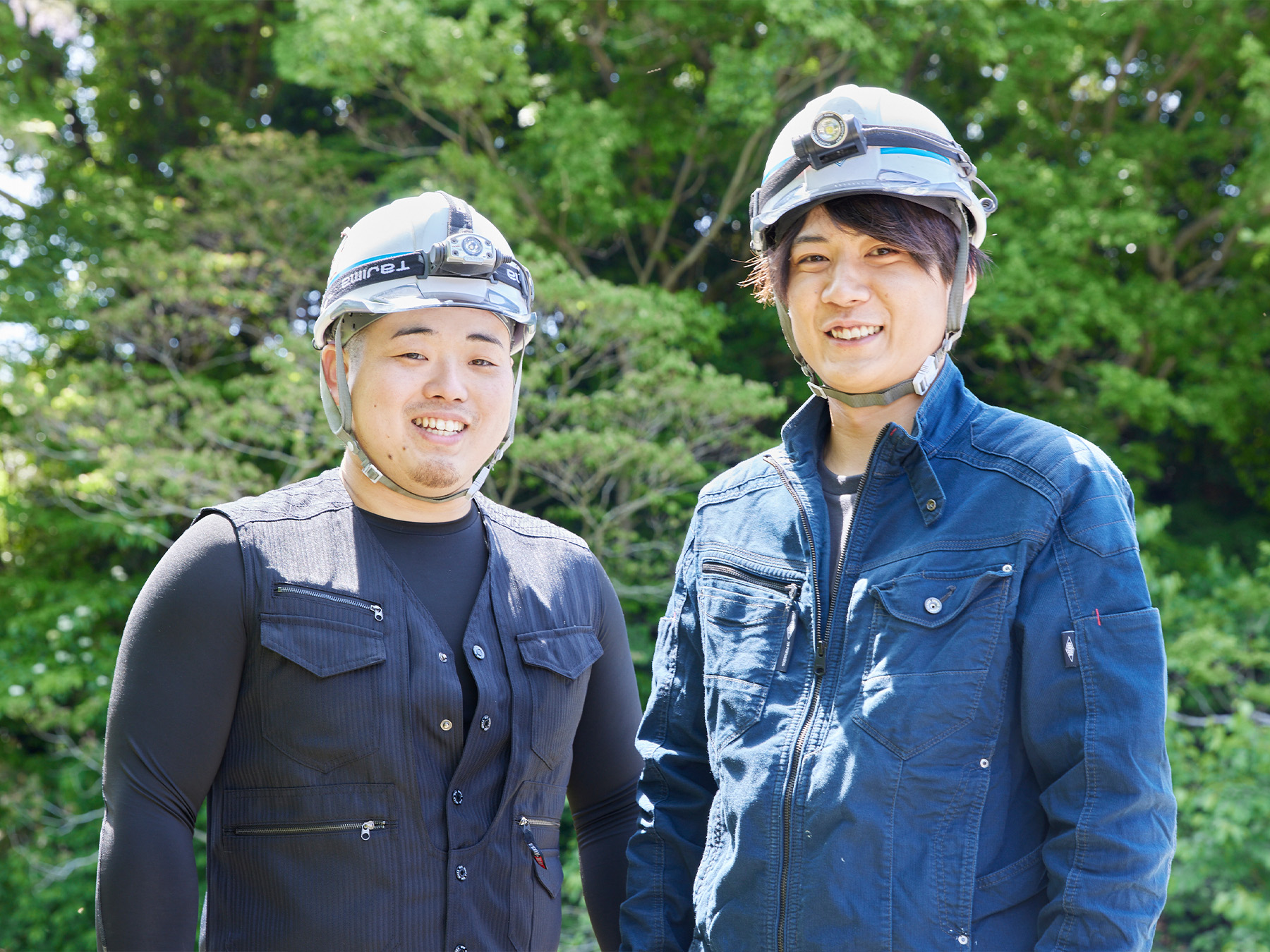 緑豊かな背景の中、笑顔で並ぶ二人の作業員のポートレート
